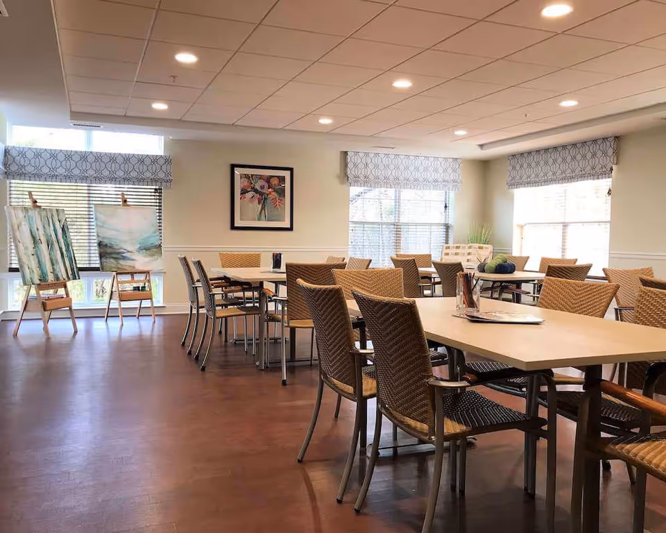 Bright common room with multiple tables and wicker-style chairs, large windows with patterned valances, and easels displaying artwork.