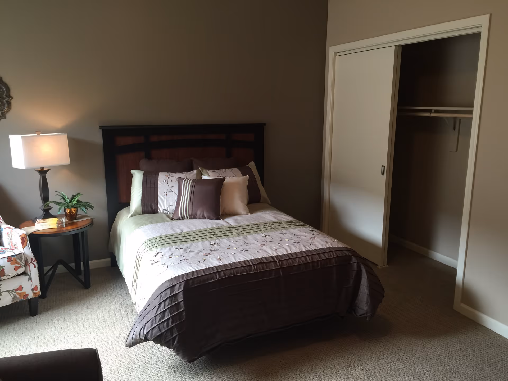 A bedroom with a neatly made bed featuring multiple pillows and a patterned bedspread. To the left of the bed is a round wooden side table with a lamp and a small potted plant. There is a floral upholstered chair partially visible on the left side. On the right side of the room, there is an open closet with sliding doors. The walls are painted beige and the floor is carpeted.