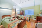 A bright and clean bedroom with two single beds, each with colorful bedspreads. There is a wooden nightstand with a lamp between the beds, a wooden wardrobe, a window with blinds, and two wooden chairs along the wall. The room has light yellow walls and wooden flooring.