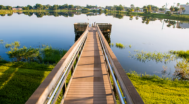 A wooden boardwalk leading to a small dock over a calm lake with grassy shoreline and trees reflected in the water.