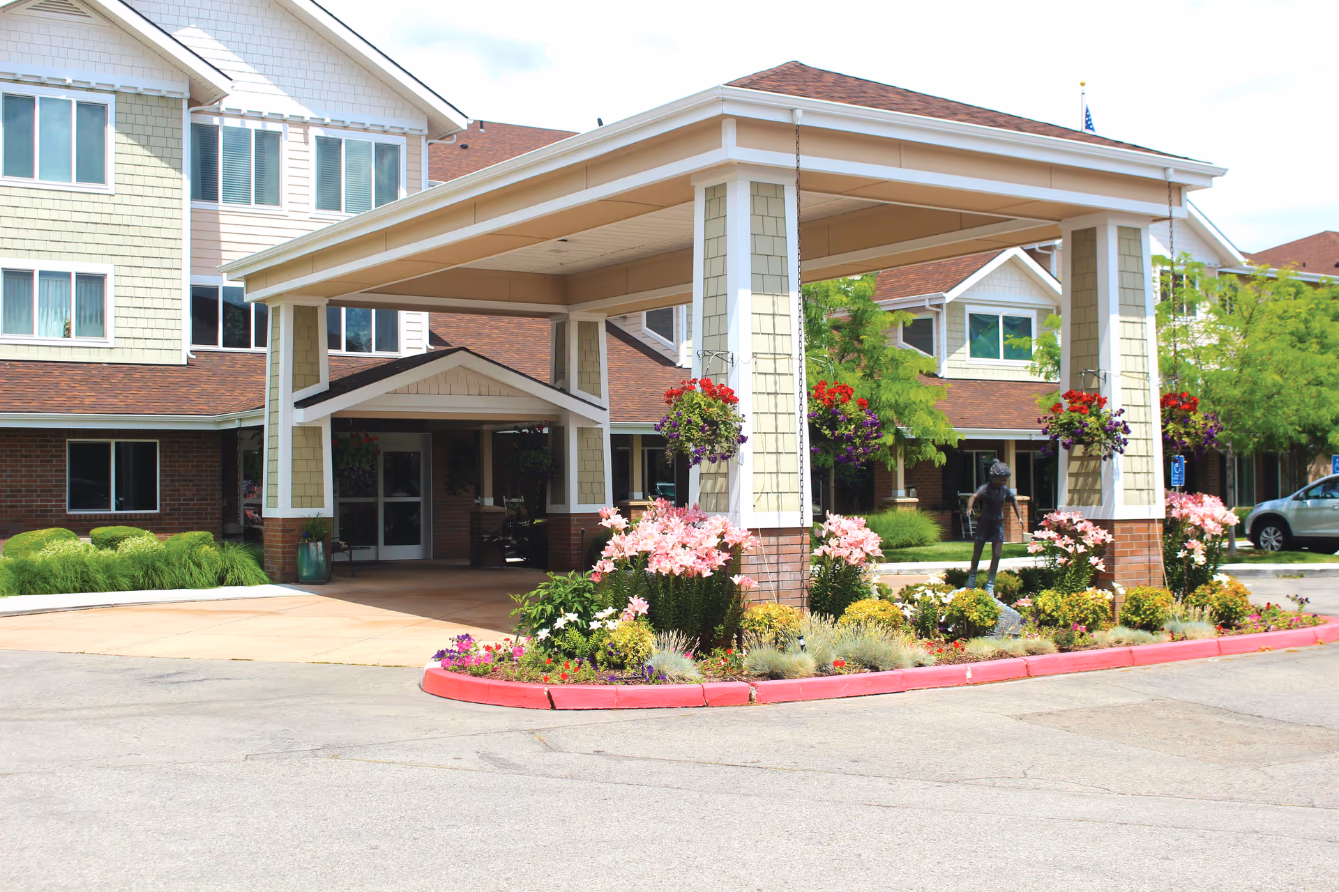 Entrance of a senior living facility with a covered drop-off area supported by columns. The area is decorated with hanging flower baskets and a landscaped garden bed with various colorful flowers. The building has multiple windows and a combination of brick and siding exterior.