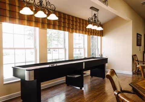 A bright room with large windows covered by striped valances, featuring a shuffleboard table in the center. The room has wooden flooring and is furnished with wooden chairs and a table partially visible on the right side. Two ceiling light fixtures with multiple bulbs hang above the shuffleboard table.