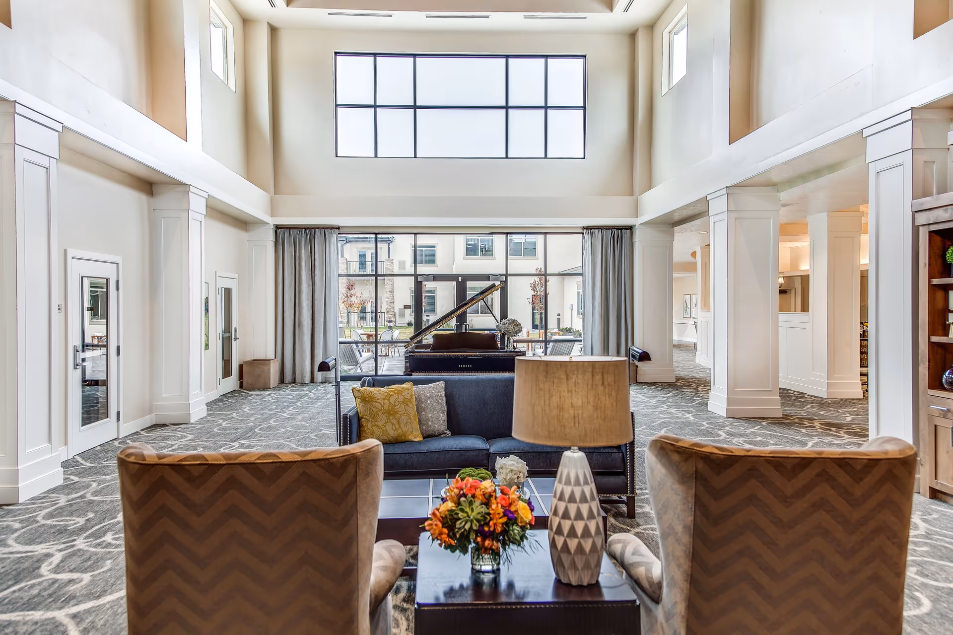 Spacious two-story common lounge with sofas and chairs facing a grand piano under large windows.