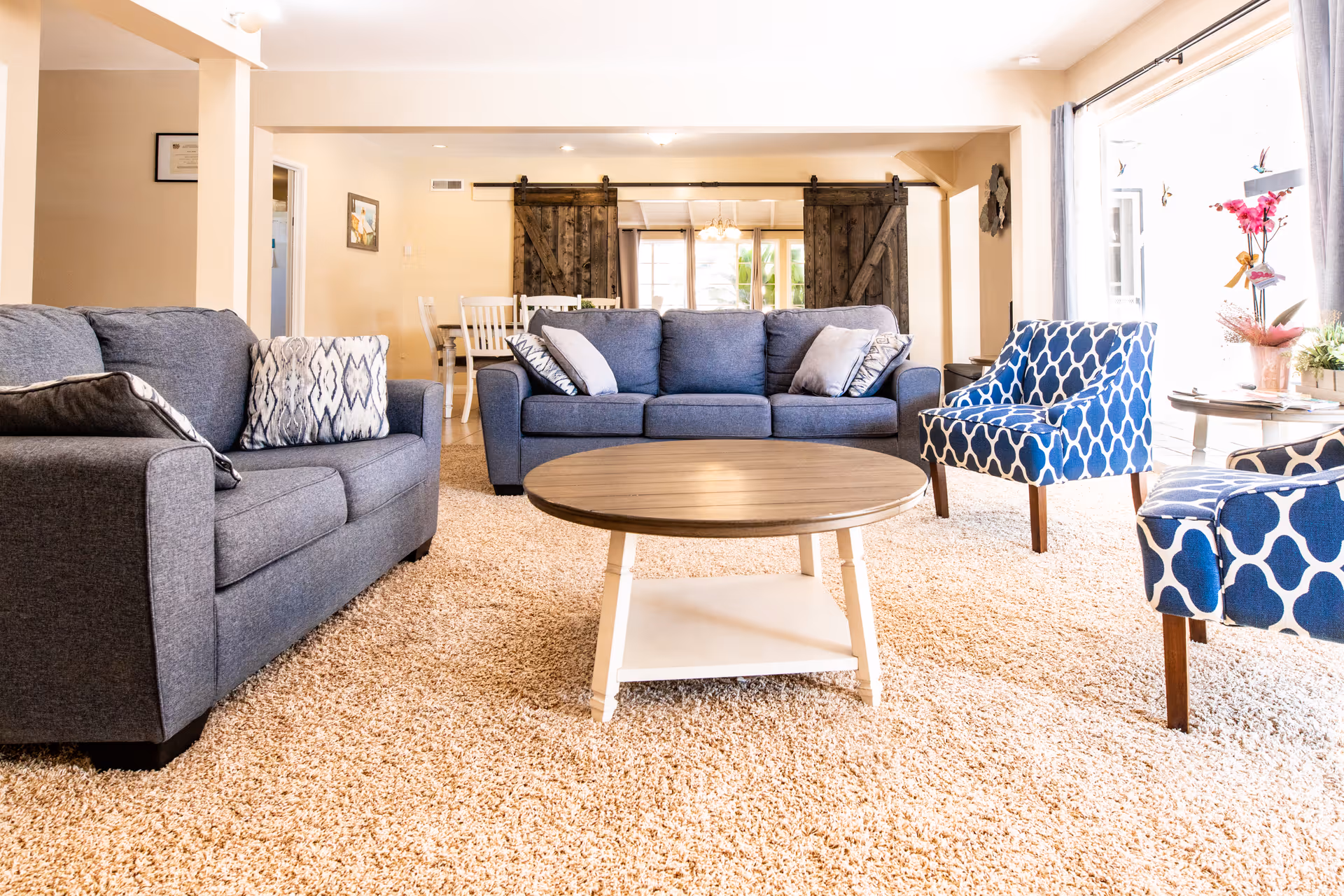 A bright and cozy living room with beige carpet, two gray sofas with patterned cushions, two blue and white patterned armchairs, and a round wooden coffee table in the center. In the background, there is a dining area with white chairs and a table, separated by rustic wooden sliding barn doors. Large windows with curtains allow natural light to fill the room.