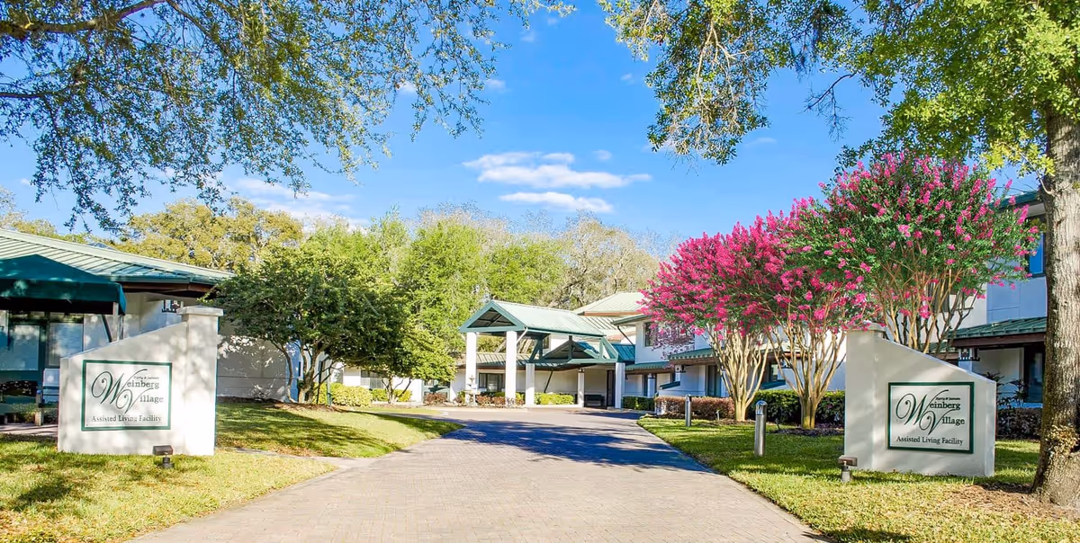 Entrance driveway to Weinberg Village Assisted Living Facility with a covered drop-off area, surrounded by green trees and vibrant pink flowering bushes under a clear blue sky.