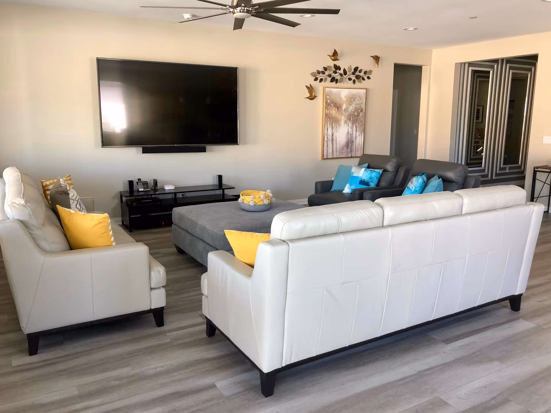 A modern living room with two white leather sofas and two gray armchairs arranged around a large gray ottoman. The sofas have yellow and gray decorative pillows, while the armchairs have blue pillows. A large flat-screen TV is mounted on the wall above a black TV stand. The room features light wood flooring, a ceiling fan, and wall art with metallic bird decorations.