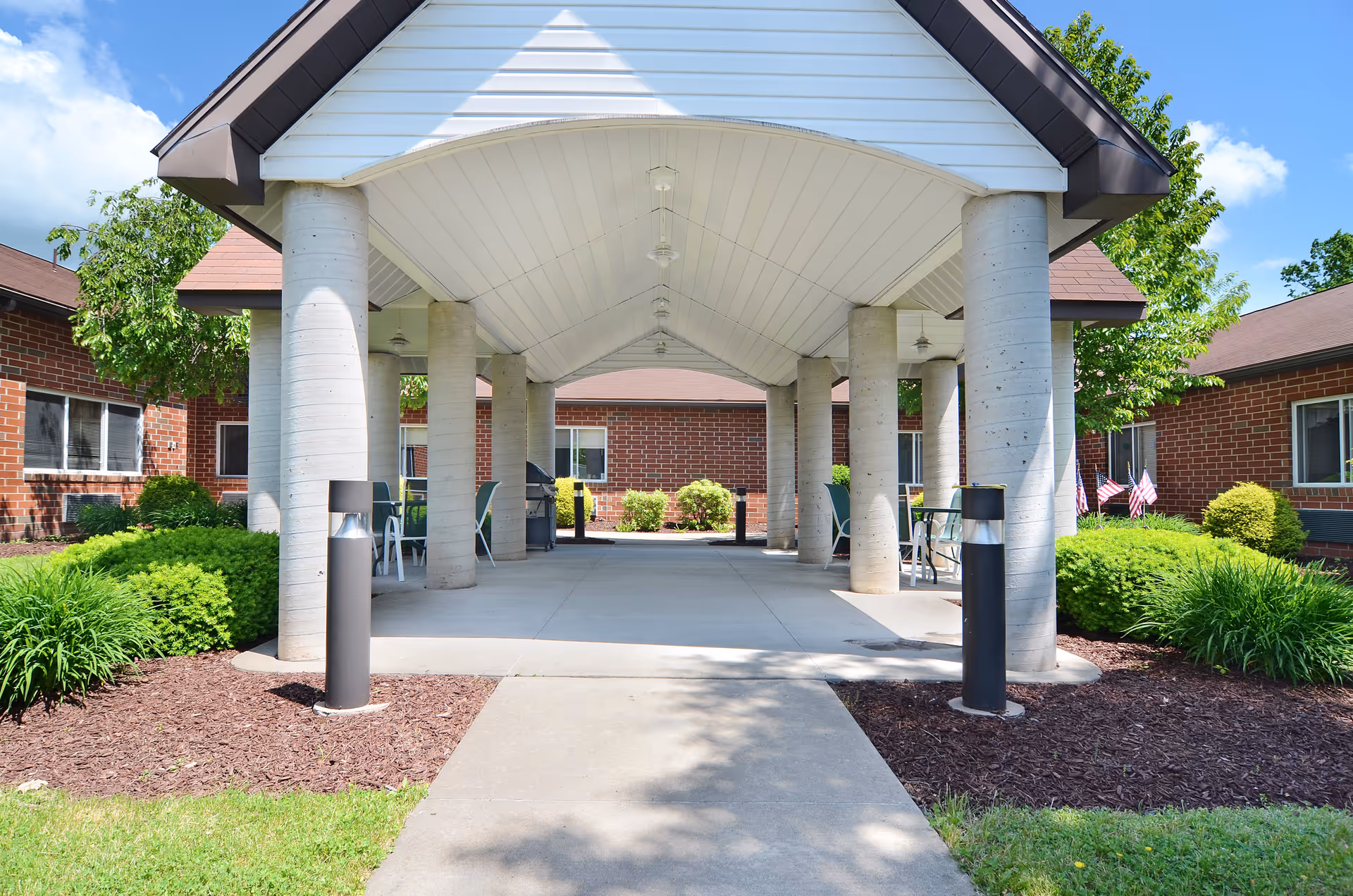 Covered outdoor walkway with large concrete pillars leading to a brick building, surrounded by green bushes and trees under a blue sky with some clouds.