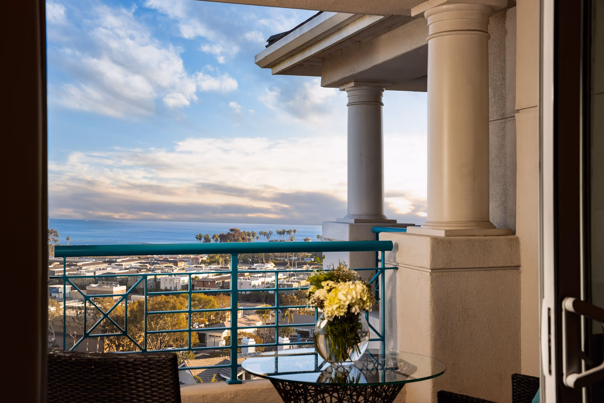 View from a balcony with a glass table holding a vase of white flowers, two wicker chairs, and a railing overlooking a residential area with trees and the ocean in the background under a partly cloudy sky.