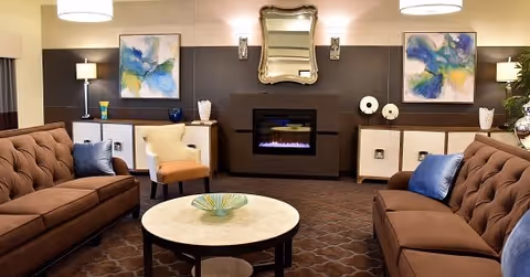 A cozy living room with two brown tufted sofas facing each other, a round coffee table in the center, and a beige armchair. The room features a modern electric fireplace with a decorative mirror above it, two abstract paintings on the wall, and soft lighting from ceiling fixtures and table lamps.