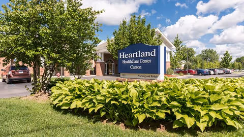 Exterior view of Heartland HealthCare Center Canton showing the building entrance, a blue sign with the facility name, surrounded by green plants and trees under a partly cloudy blue sky. Several cars are parked in the parking lot to the right.
