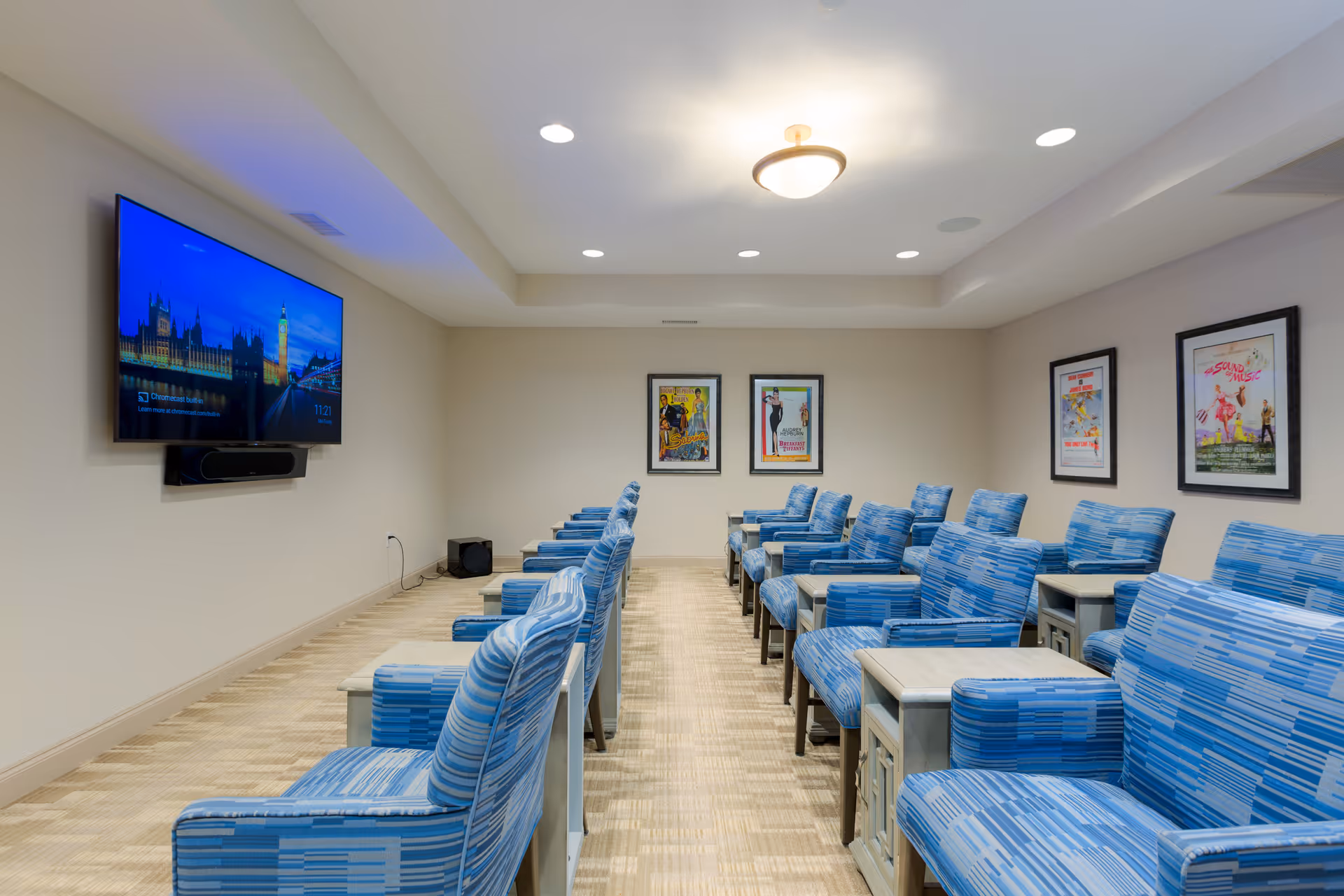 A small theater room with two rows of blue patterned armchairs facing a wall-mounted flat screen TV. The room has beige walls, a light-colored carpet, recessed ceiling lights, and framed movie posters on the far wall.