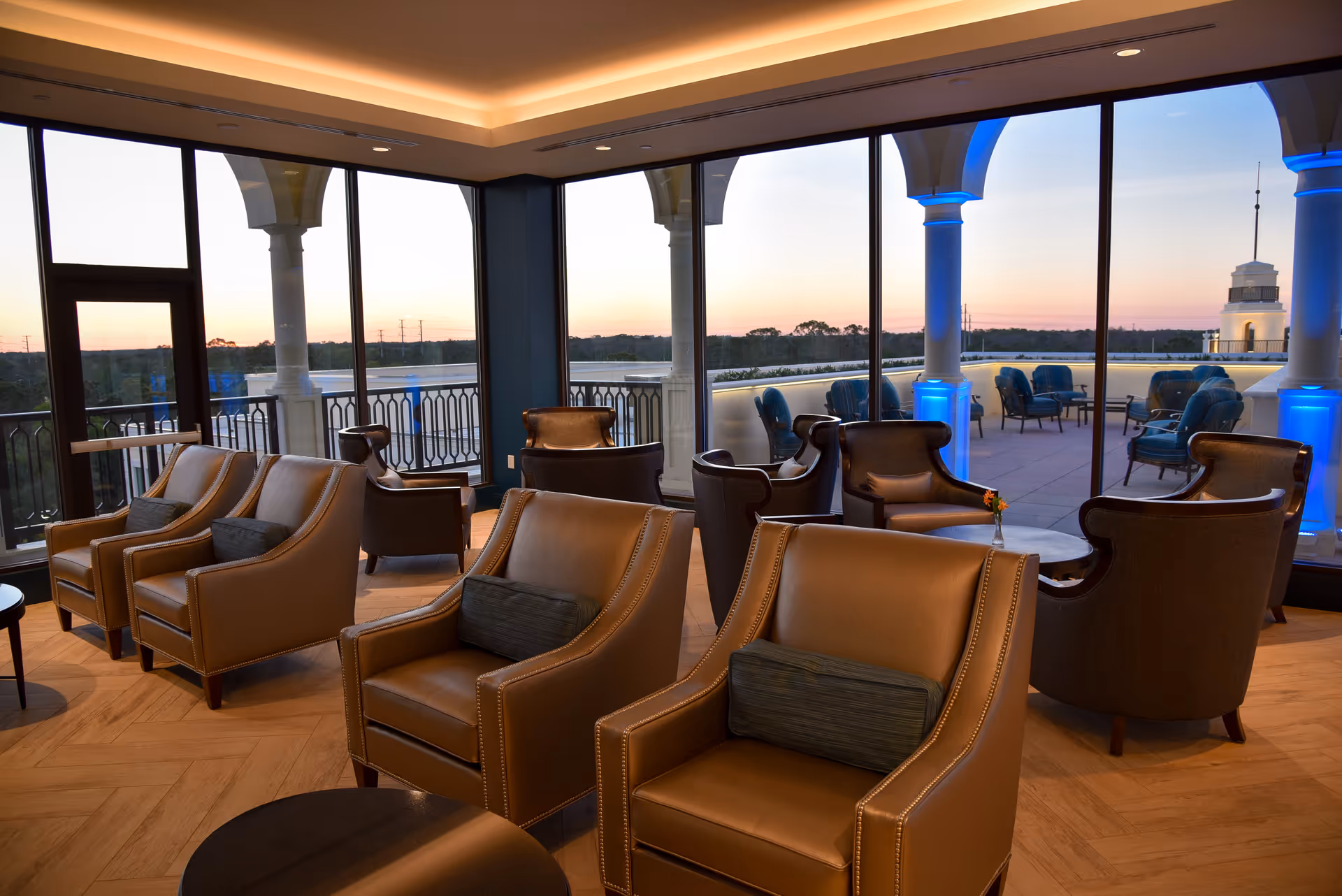 A cozy indoor lounge area with multiple leather armchairs and small tables, large floor-to-ceiling windows offering a view of an outdoor patio with additional seating and columns illuminated with blue lights, during sunset.