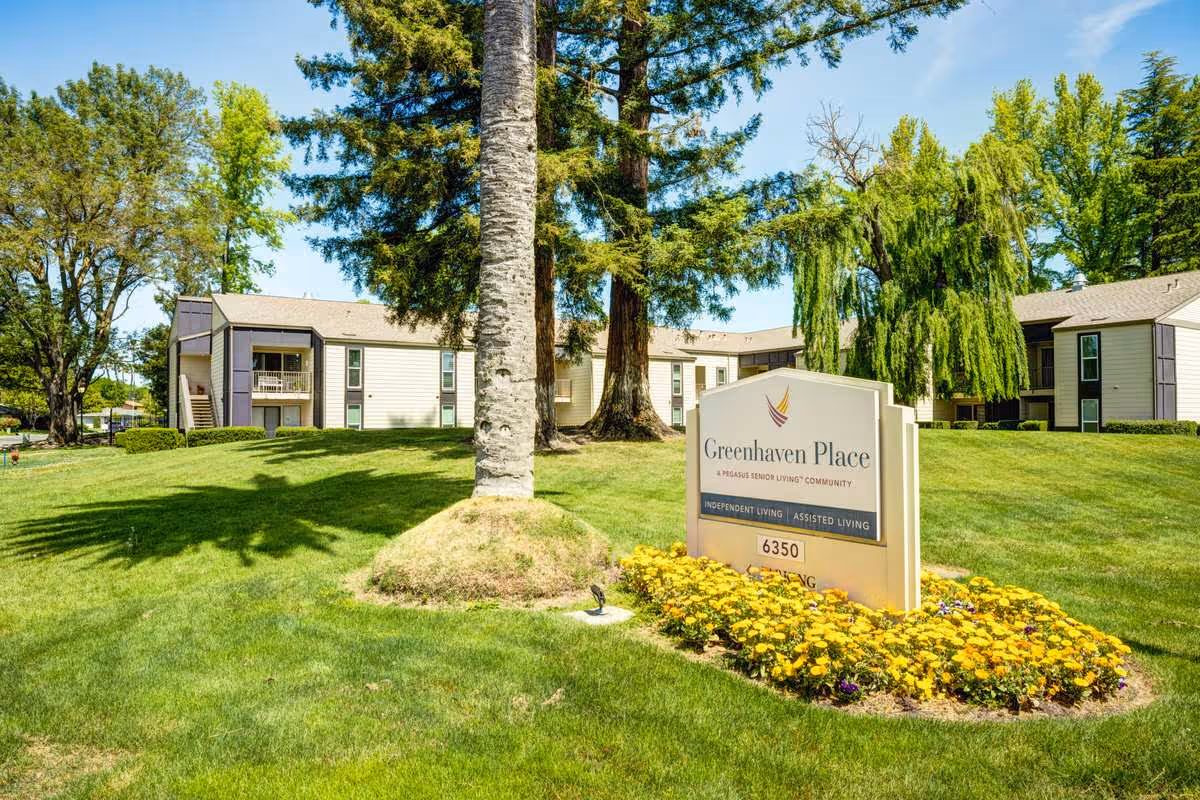 Exterior view of Greenhaven Place senior living community with a large green lawn, trees, and a sign displaying the facility's name and services including independent living and assisted living.