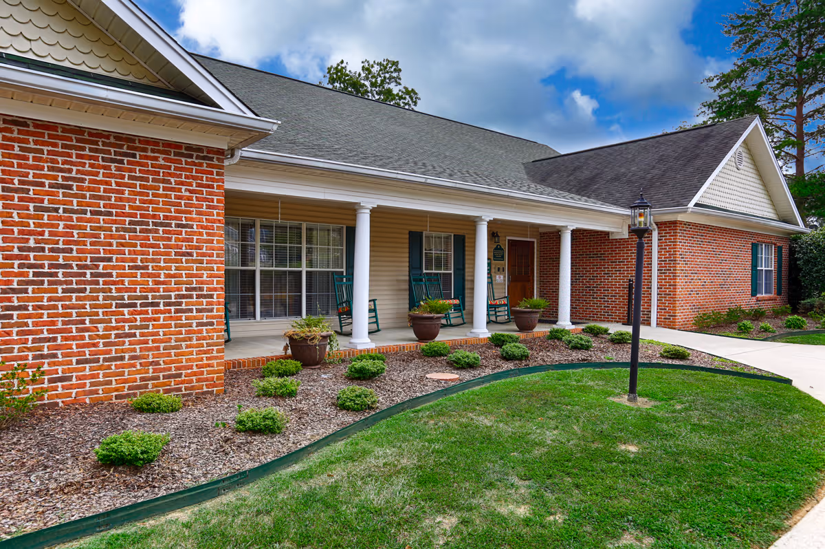 Exterior view of a single-story brick building with a covered porch featuring white columns, green rocking chairs, potted plants, and a lamp post on a grassy lawn under a partly cloudy sky.
