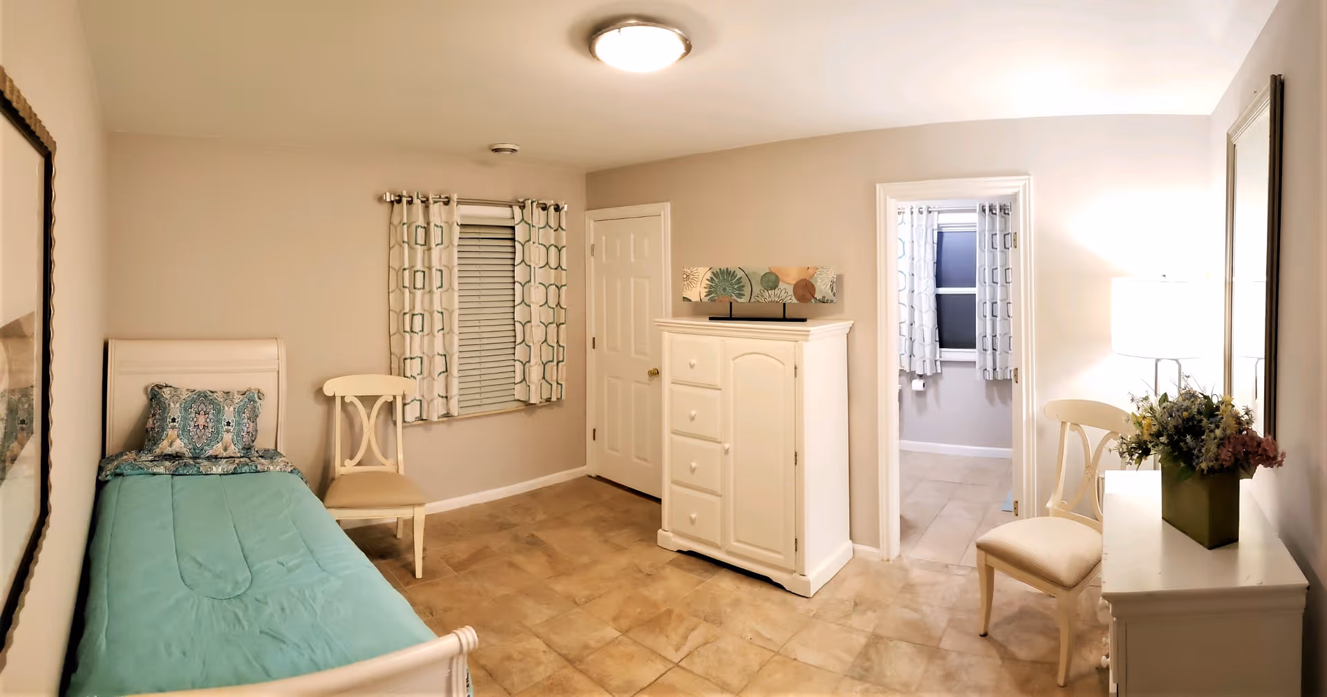 Bright furnished bedroom with a twin bed, white dresser, chairs, a lamp, and a doorway leading to an adjacent room.