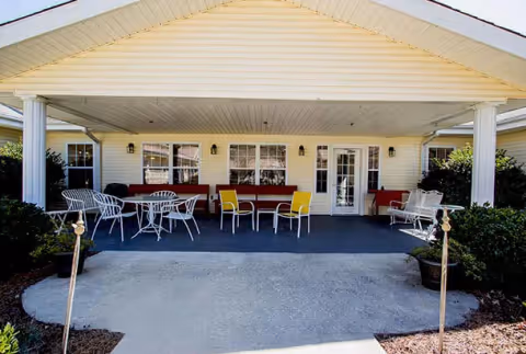 Covered front porch/entrance with tables, chairs, benches, columns, and potted plants at a senior living facility.
