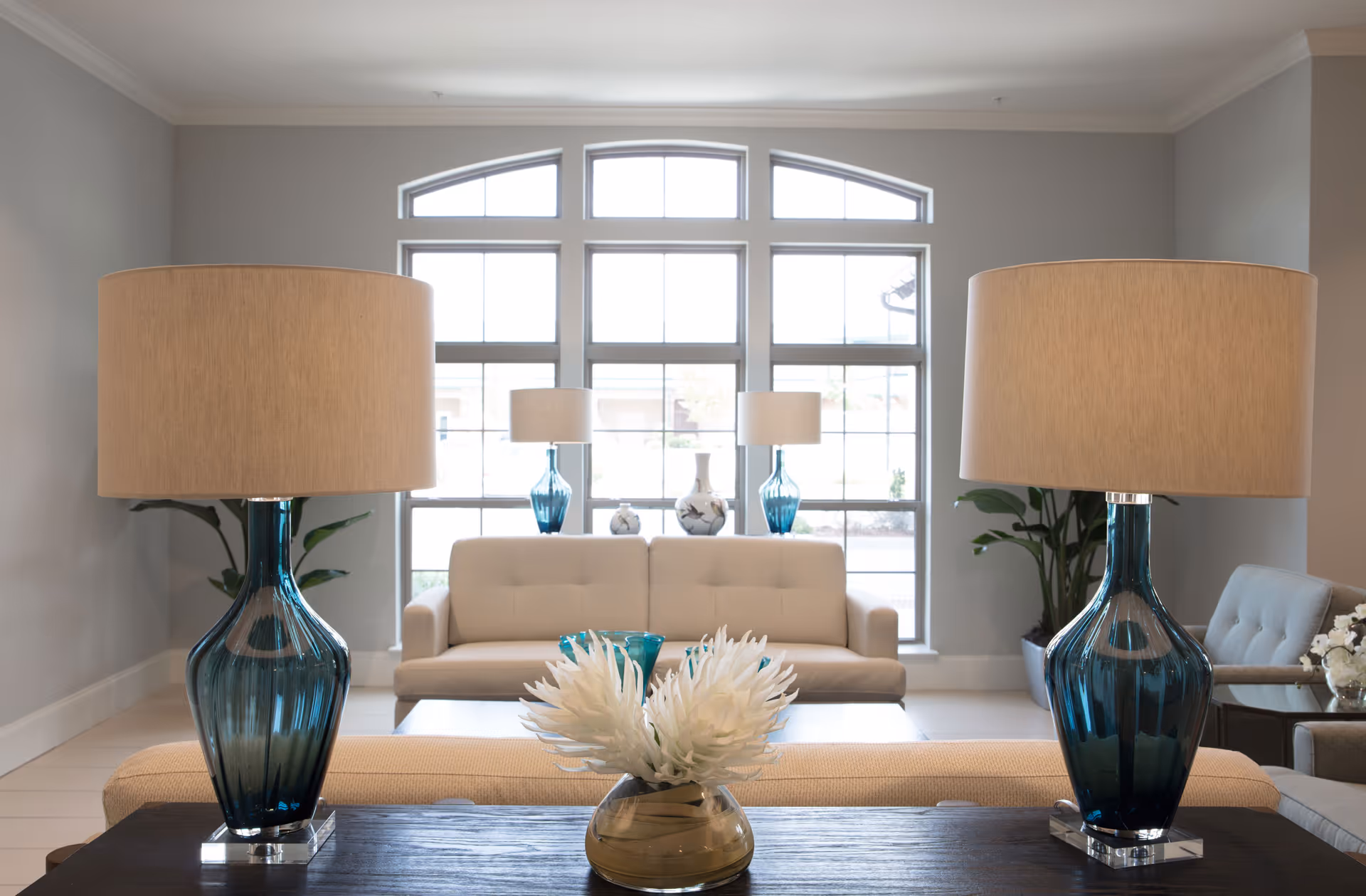 A bright living room with large windows letting in natural light. The room features a beige sofa, two blue glass table lamps with beige shades on a dark wooden table, a beige cushioned bench, and a vase with white flowers. There are also two blue glass vases and a white decorative vase on a table in front of the sofa, with green plants in the corners of the room.