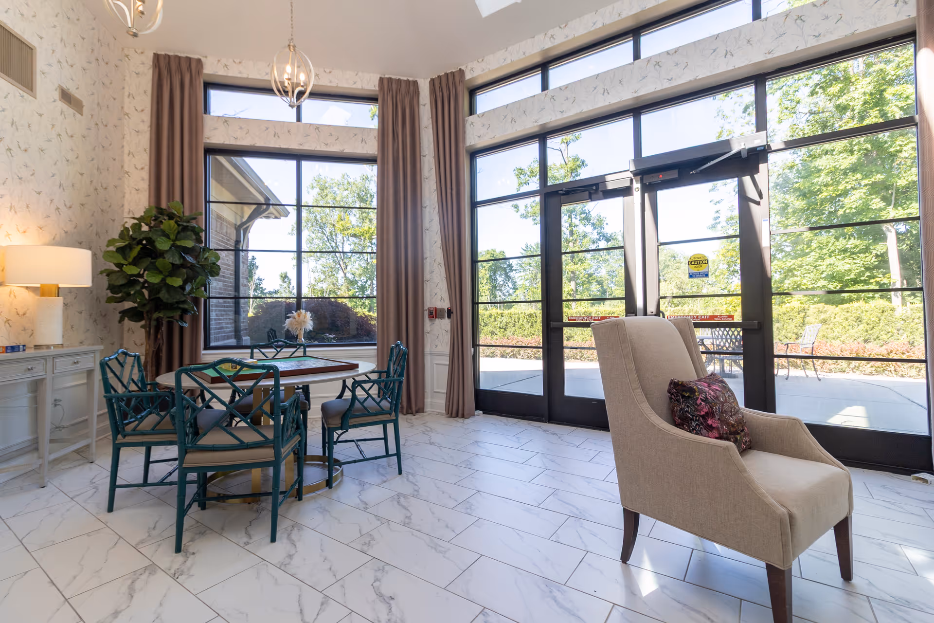 Bright senior living common area with large windows and glass doors, a round table with green chairs, and a beige armchair.