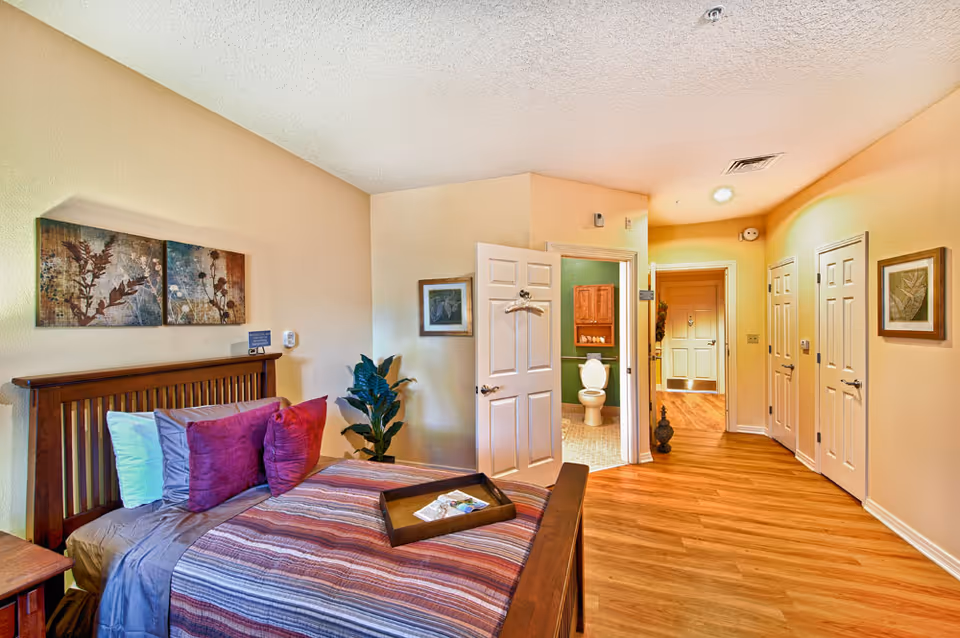 Furnished bedroom with a wooden bed, colorful pillows and blanket, open door showing a bathroom and hallway.