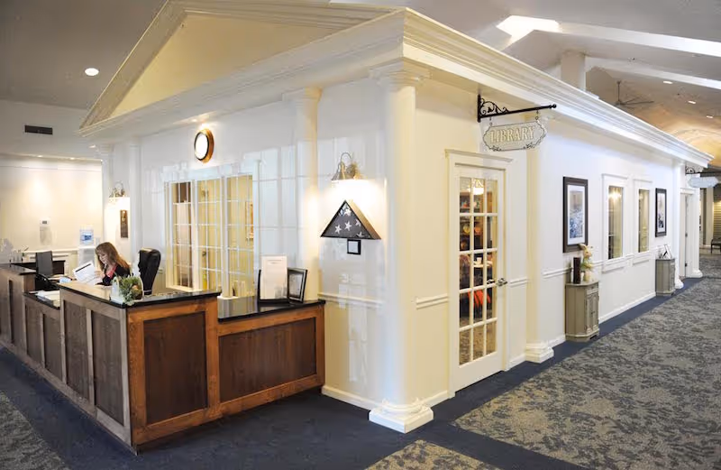 Interior view of a senior living facility reception area with a wooden front desk where a woman is seated working. Adjacent to the desk is a small building-like structure labeled 'Library' with a glass door and windows. The area is well-lit with wall sconces and has carpeted flooring.