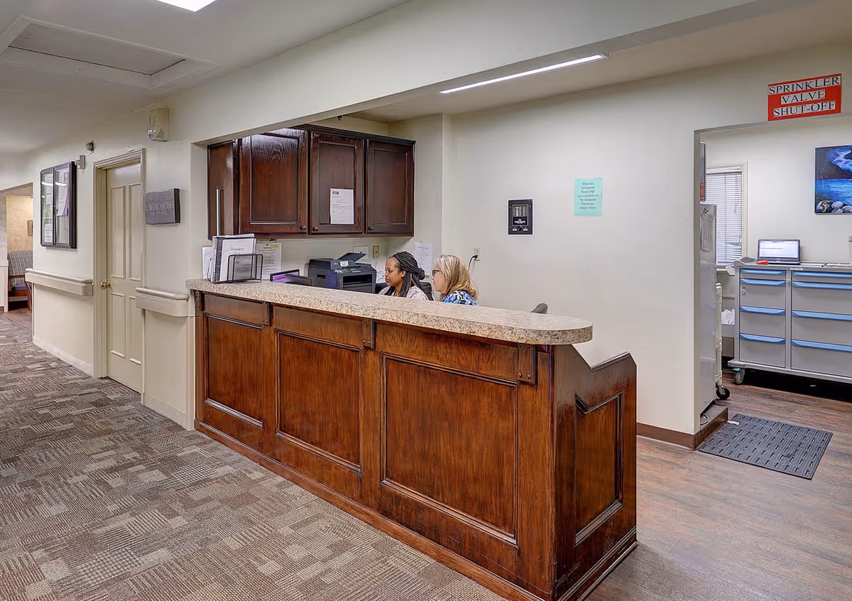Reception area inside a senior living facility with a wooden front desk and two staff members working behind it. The hallway has carpeted flooring and light-colored walls with handrails. There is a door and framed pictures on the wall to the left, and an open doorway to the right leading to a room with cabinets and a computer.