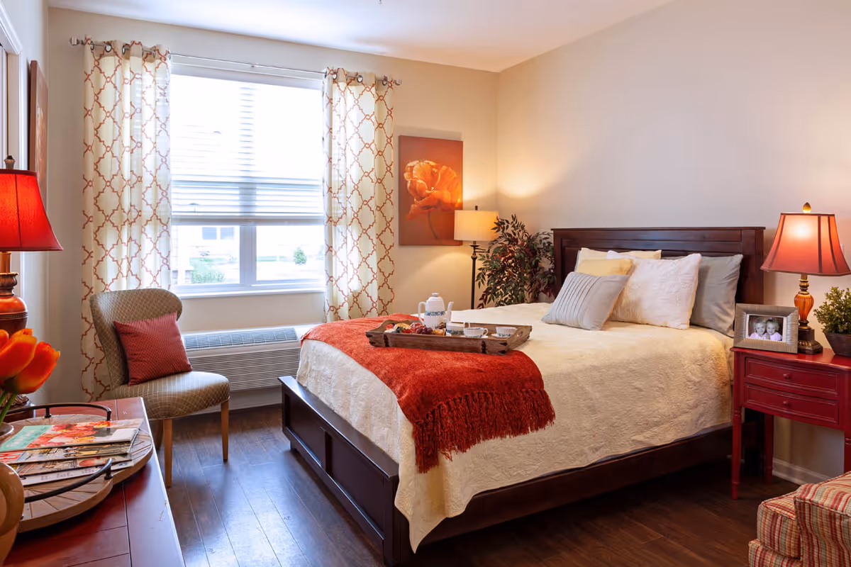 A cozy bedroom with a wooden bed frame, cream-colored bedding, and a red throw blanket. There is a tray with a teapot and cups on the bed. The room has two red lamps on either side of the bed, a chair with a red cushion near a window with patterned curtains, a small red nightstand with a framed photo, and a potted plant in the corner.