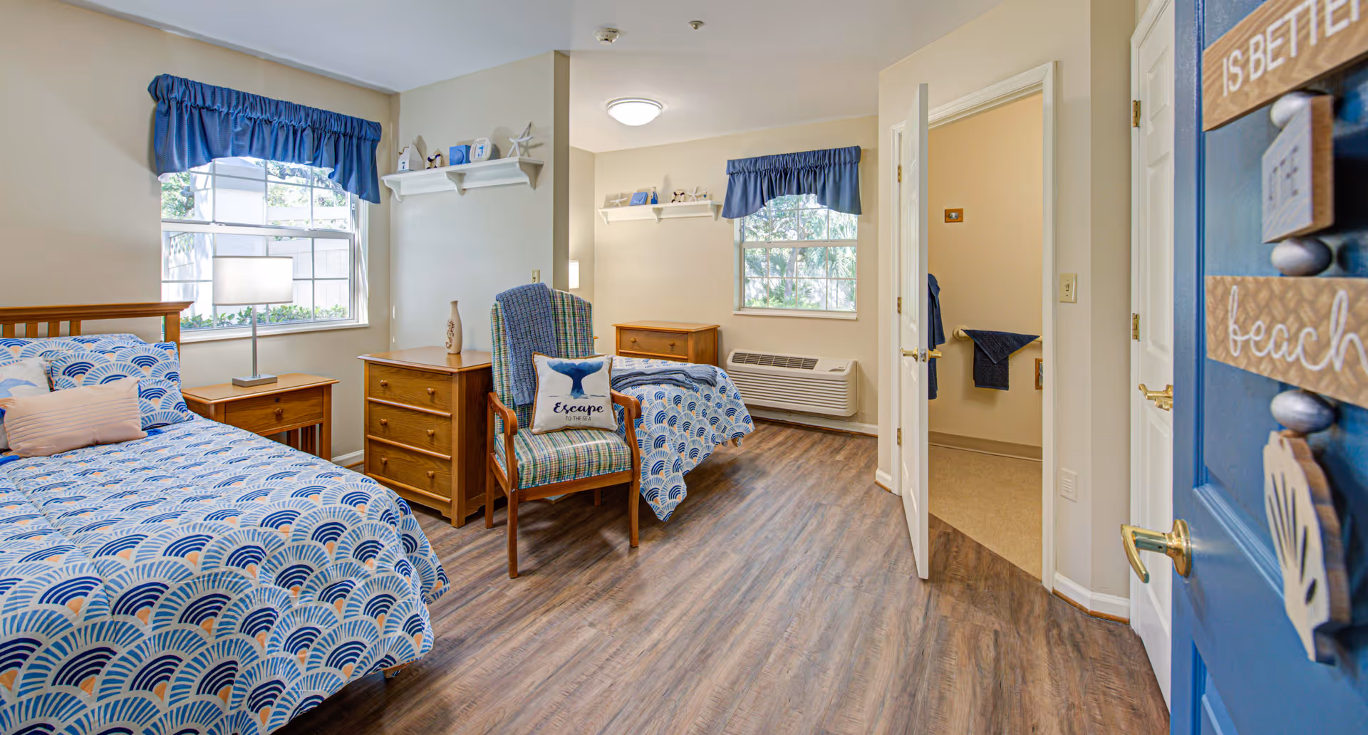 A bright and cozy bedroom with two twin beds covered in blue and white patterned bedding. There is a wooden nightstand with a lamp between the beds, a wooden dresser, and a cushioned chair with a pillow that says 'Escape'. The room has two windows with blue valances, light-colored walls, and wood flooring. A door is open, leading to a bathroom with visible towels. The entrance door is blue with beach-themed decorations.