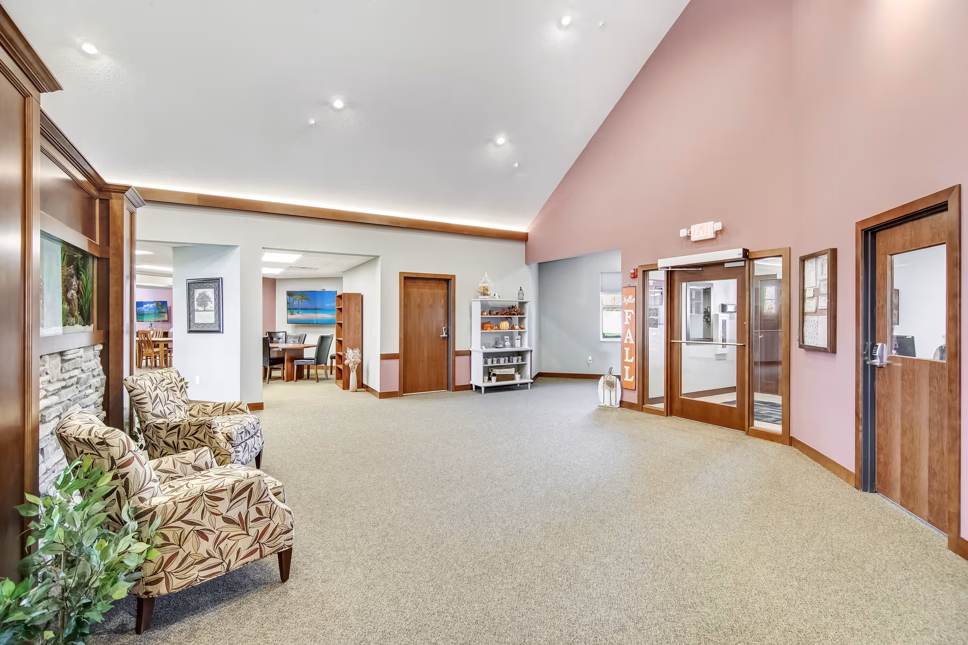 A spacious interior common area with beige carpet and light purple walls. Two patterned armchairs are placed near a stone fireplace on the left. There is a white shelving unit with decorative items and a sign that says 'Hello FALL' near the glass double doors on the right. A dining area with a table and chairs is visible through an open doorway in the background.