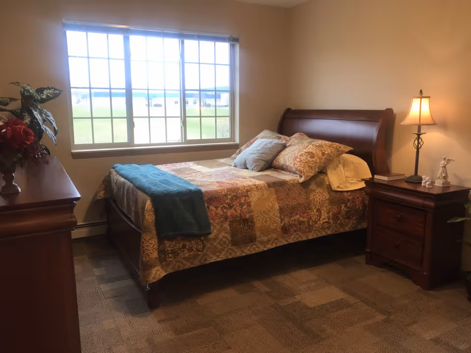 A cozy bedroom with a large window letting in natural light. The room features a wooden bed with a patterned quilt and multiple pillows, a wooden nightstand with a lamp and decorative items, and a wooden dresser with a vase of flowers. The carpeted floor has a checkered pattern.