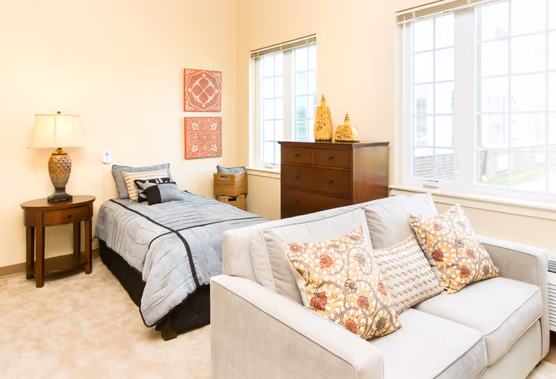 A bright and cozy bedroom featuring a single bed with gray bedding and decorative pillows, a wooden nightstand with a lamp, a wooden dresser with decorative items on top, and a light-colored sofa with patterned throw pillows. Two large windows let in natural light, and two red decorative wall hangings are above the bed.