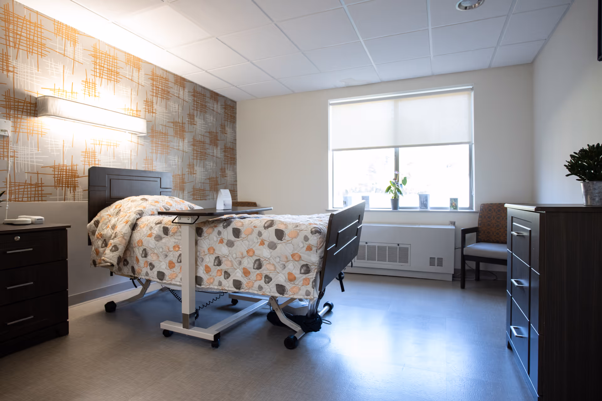 A bright and clean bedroom in a senior living facility with a hospital-style adjustable bed covered with a patterned quilt. The room has a window with a roller shade, a small table over the bed, a wooden dresser, a nightstand with a telephone, a chair, and a potted plant on the dresser. The wall behind the bed features a decorative wallpaper with an abstract pattern.