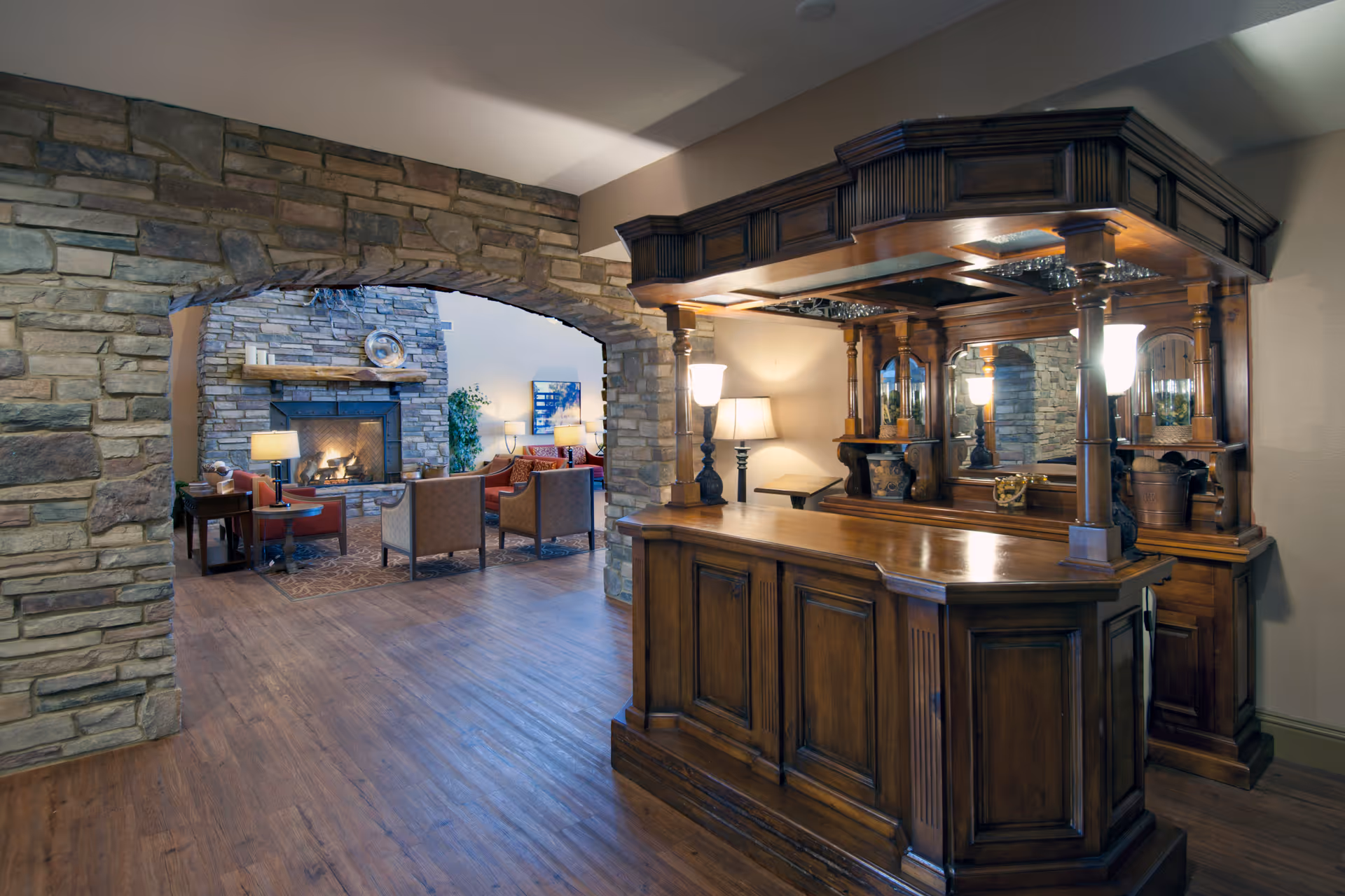 Interior view of a senior living facility featuring a wooden bar with lamps and glassware on the right, and a cozy seating area with armchairs and a stone fireplace visible through a stone archway on the left.