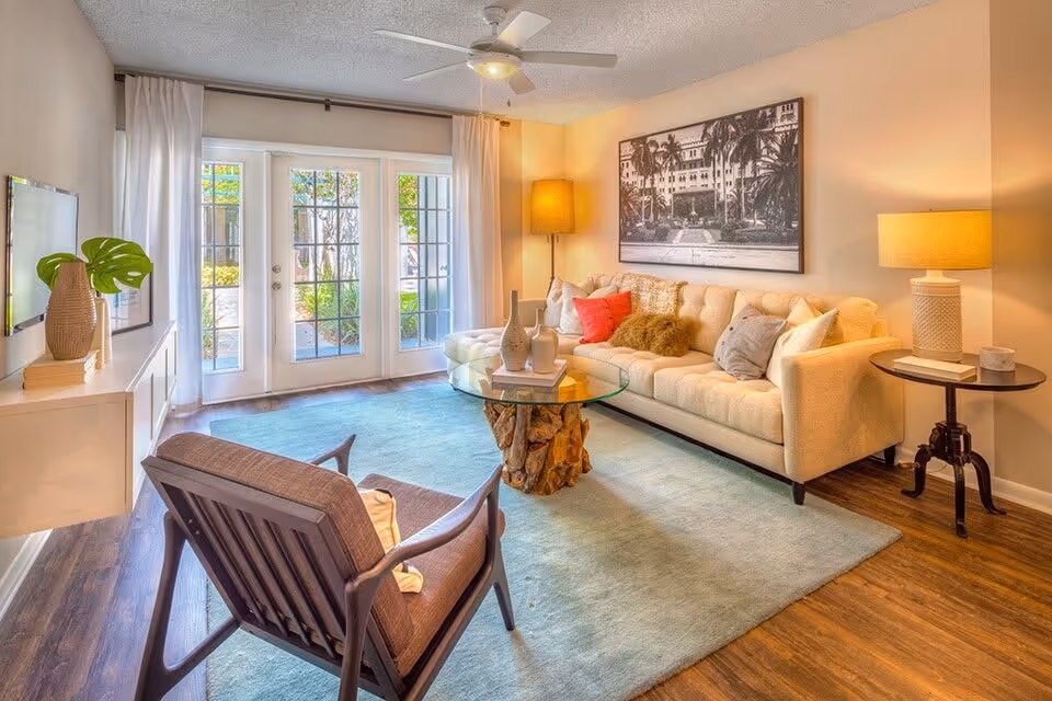 Sunny living room with a beige tufted sofa, armchair, glass-top driftwood coffee table, area rug, and French doors to a patio.