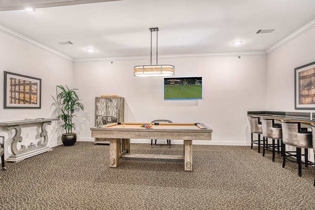 A recreational room with a pool table in the center, a flat-screen TV mounted on the wall showing a sports game, a tall potted plant, framed artwork on the walls, a decorative cabinet, a console table, and a bar counter with four high chairs.