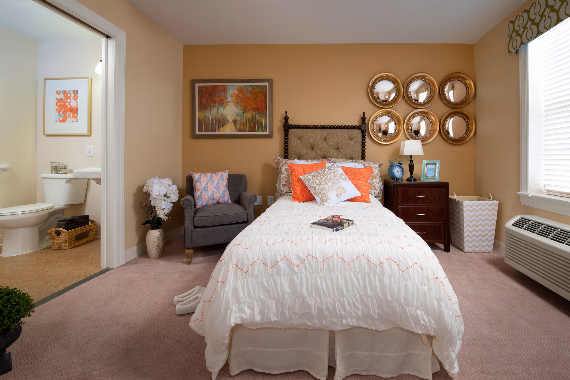 A cozy bedroom with a neatly made bed featuring white and orange pillows and a white comforter. To the left of the bed is a gray armchair with a decorative pillow and a vase with white flowers. Above the bed is a framed painting of a tree-lined path and a wall decoration of six round mirrors arranged in two rows. To the right of the bed is a wooden nightstand with a lamp, a blue alarm clock, and a picture frame. A laundry basket is placed next to the nightstand. The room has beige walls and carpeted floor. A window with blinds and a patterned valance is on the right wall. An air conditioning unit is below the window. A bathroom with a toilet and sink is visible through an open door on the left side of the image.