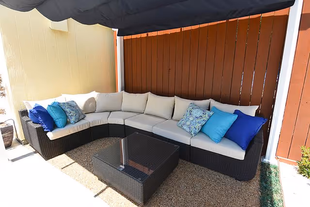 Outdoor patio with a curved rattan sectional sofa topped with beige cushions and blue decorative pillows and a glass-top coffee table under a canopy.
