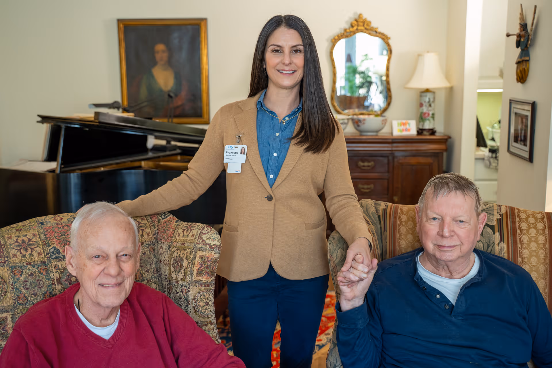 A woman standing between two elderly men seated in armchairs in a cozy living room. The woman is smiling and holding hands with one of the men. Behind them is a grand piano, a framed portrait, a wooden dresser with a decorative mirror, a lamp, and some small decorative items.