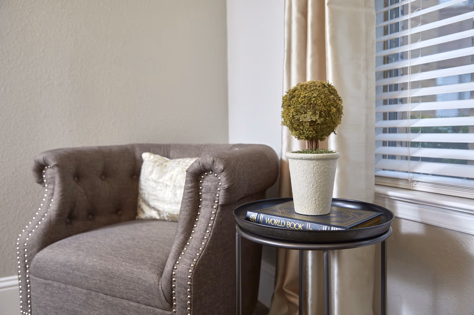 Upholstered gray armchair beside a round side table holding a potted topiary and a World Book book by a window with blinds.
