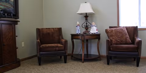 Two brown upholstered armchairs with nailhead trim are positioned on either side of a small wooden side table. The table holds a lamp with a white shade and two small decorative figurines. The room has beige carpet and light-colored walls with a window partially visible on the right side.