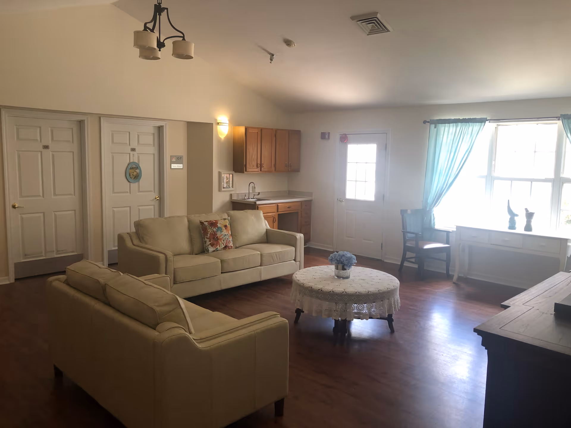 A cozy living room area with two beige sofas, a round coffee table covered with a lace tablecloth, a small kitchenette with wooden cabinets and a sink, a door, and a window with light blue curtains letting in natural light. There is a chair and a white desk near the window, and two closed doors on the left side of the room.