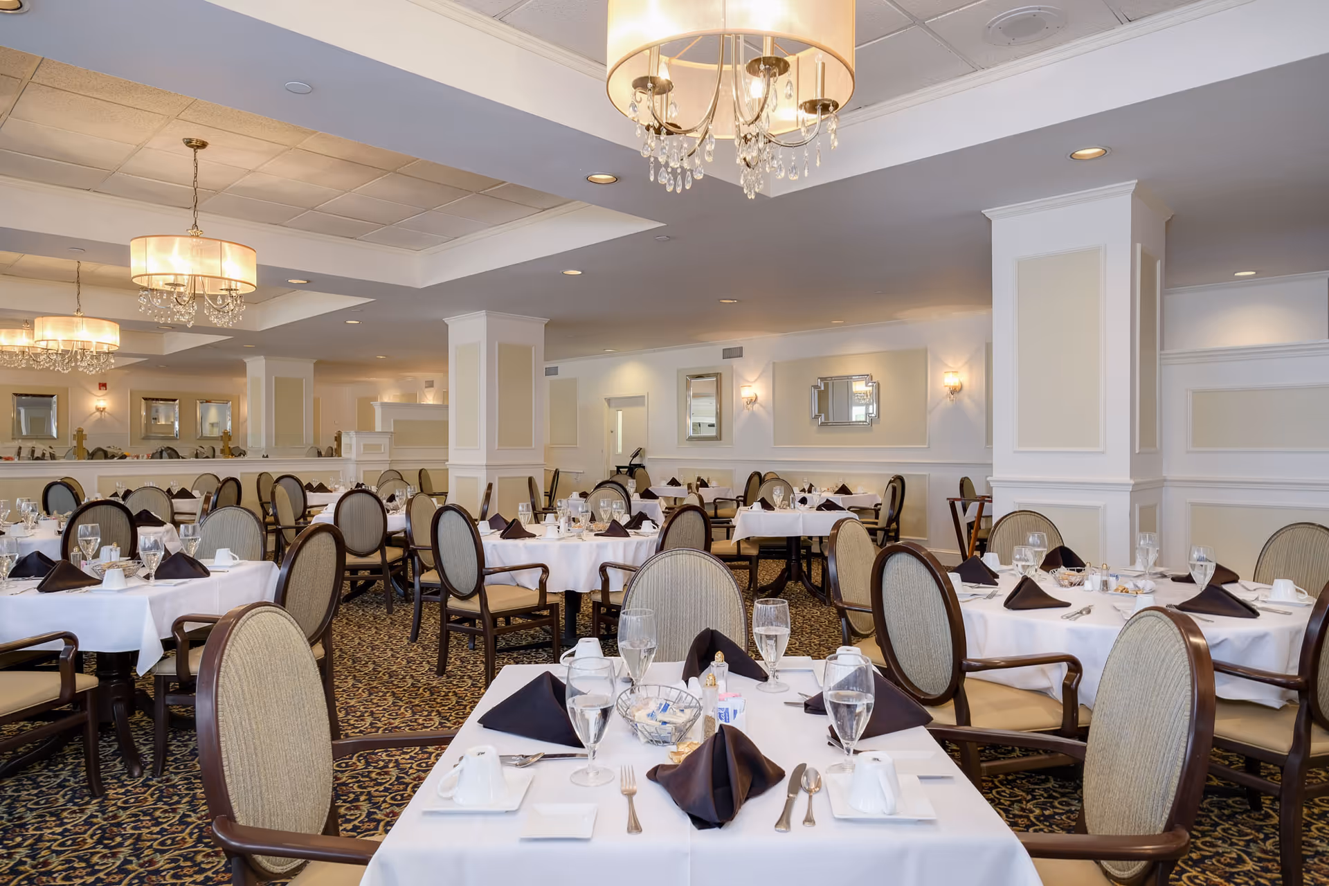 Elegant dining room with multiple round tables covered in white tablecloths, each set with glassware, silverware, black folded napkins, and small condiment containers. The room features beige walls with white trim, decorative mirrors, patterned carpet, and hanging chandeliers with crystal accents.