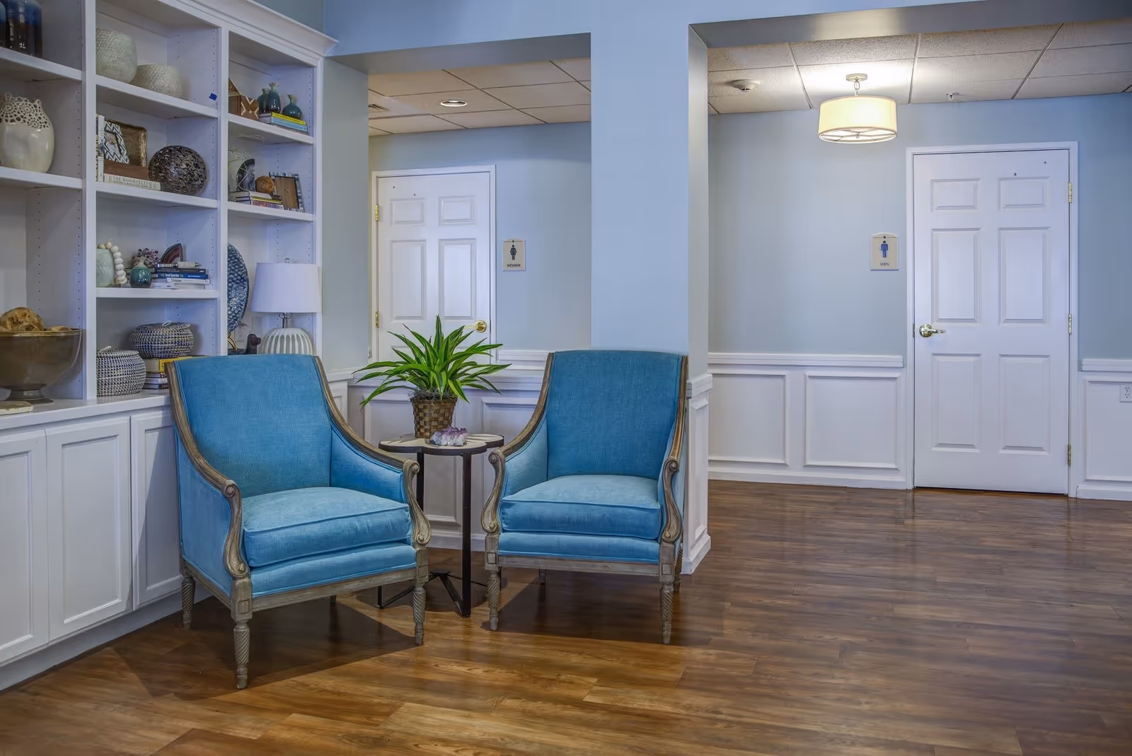 A cozy seating area in a senior living facility with two blue upholstered armchairs positioned around a small round table holding a potted plant. Behind the chairs is a built-in white shelving unit filled with decorative items and books. The walls are painted light blue with white wainscoting, and there are two white doors in the background, each marked with restroom signs. The floor is wood with a warm finish, and a ceiling light fixture is visible.