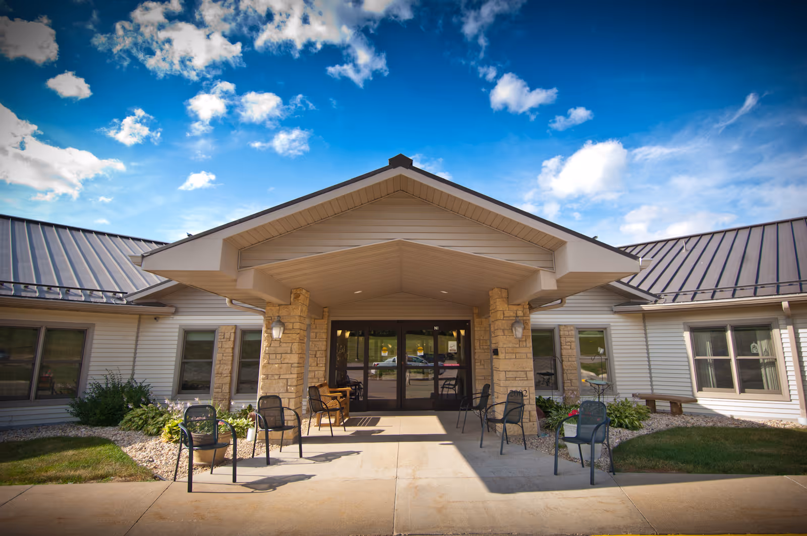 Front exterior view of Upland Hills Nursing And Rehab Center showing a covered entrance with stone pillars, several chairs arranged outside, and a clear blue sky with scattered clouds.
