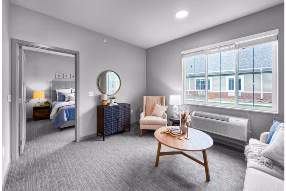 Bright furnished living room with a round coffee table, armchair, console table and large window, with a bedroom visible through an open doorway.
