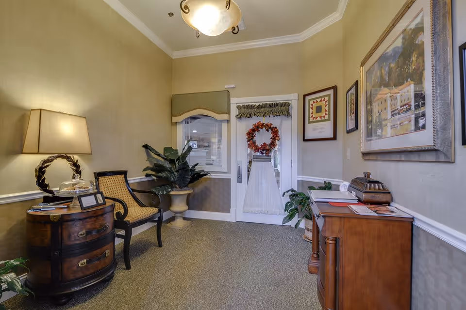 Reception seating area with a chair, lamp, side tables, potted plants, framed artwork, and a wreath-adorned door.