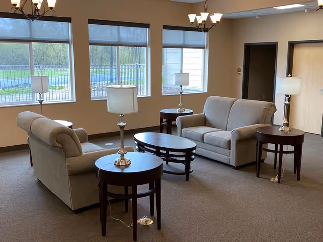 A well-lit living room area with two beige sofas facing each other, three wooden side tables each with a lamp, and a wooden coffee table in the center. Large windows with roller shades allow natural light to enter the room, and the walls are painted a neutral color.