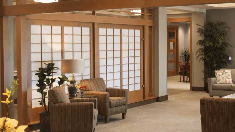 A cozy interior lounge area featuring two patterned armchairs with a small table and lamp between them, set against a backdrop of wooden framed shoji-style sliding doors. There are potted plants and additional seating visible in the background, creating a warm and inviting atmosphere.