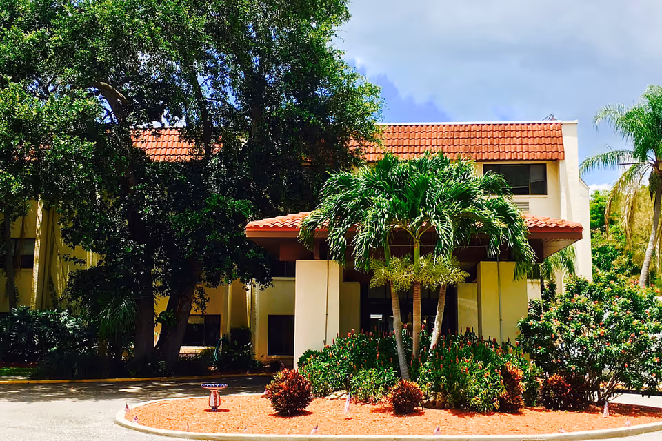 Front exterior view of a building with a red tile roof, surrounded by lush green trees and shrubs, with a circular driveway and landscaped garden in front.