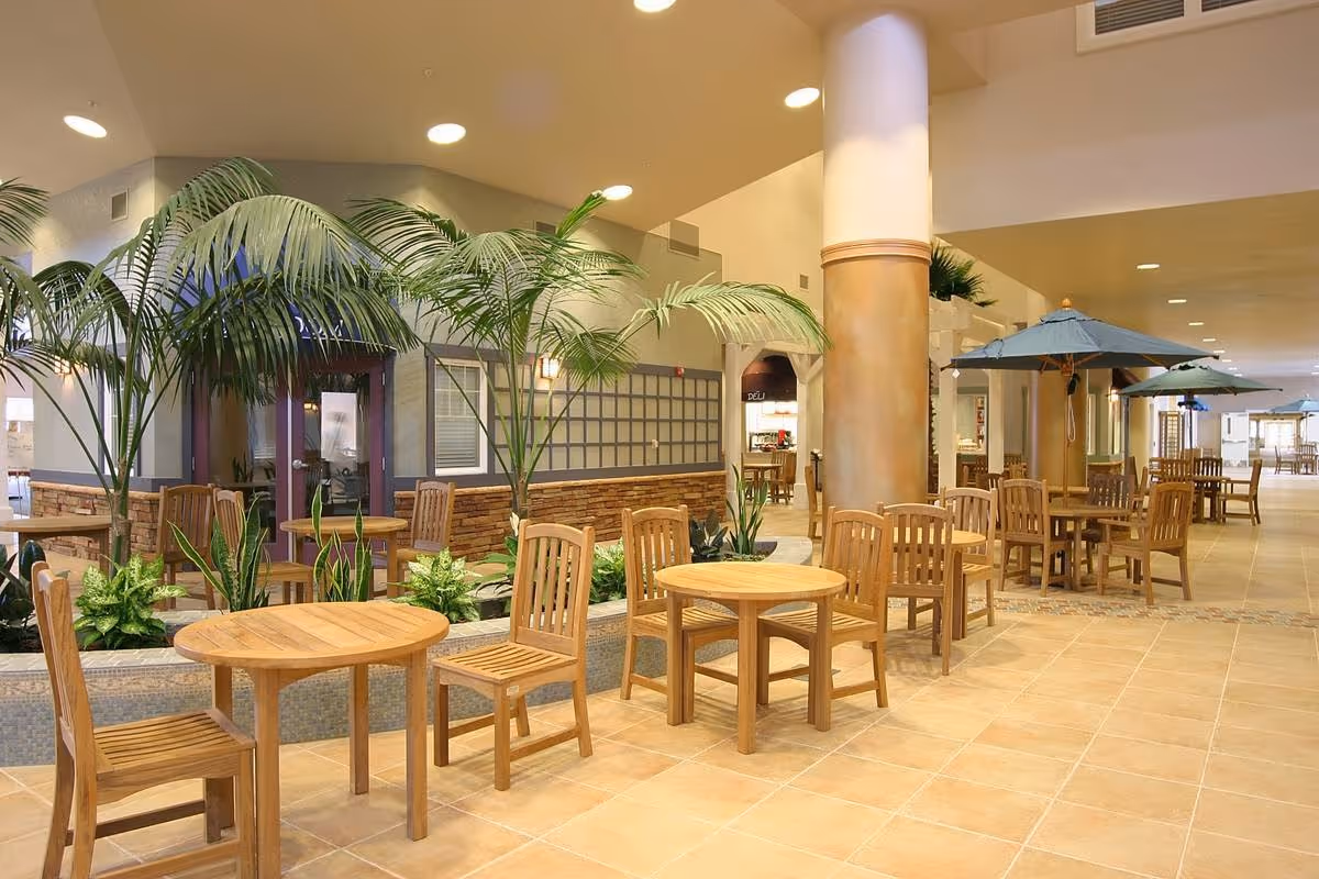 Indoor seating area with multiple wooden tables and chairs arranged along a tiled floor. Several large potted plants and palm trees are placed around the space. There are large columns and umbrellas over some tables, with a well-lit, spacious interior in the background.