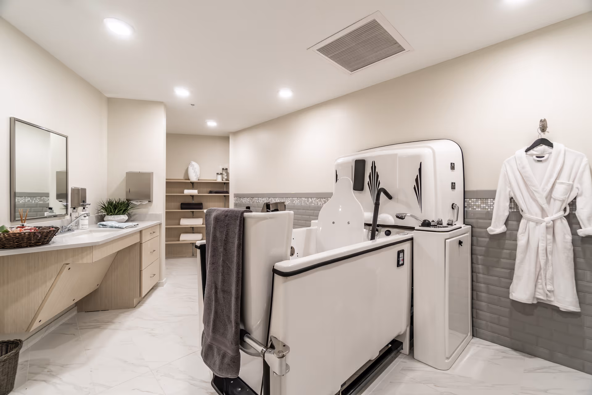 A spacious, well-lit bathroom with a large accessible bathtub equipped with safety handles and controls. A white bathrobe hangs on the wall to the right. On the left side, there is a long countertop with a sink, a mirror, a basket with toiletries, and a paper towel dispenser. Shelves with neatly folded towels and other items are visible in the background. The floor is tiled in white, and the walls are painted in light neutral tones with a decorative tile border.